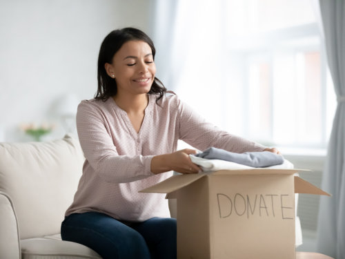 Happy African American young woman sit on couch stuck clothes in donation box at home